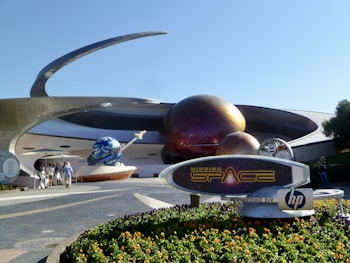 A futuristic attraction featuring large, spherical structures with a metallic appearance, resembling planets with rings. A sign in the foreground reads 'Mission: SPACE' with a logo. Visitors walk towards the entrance under a sunny sky.