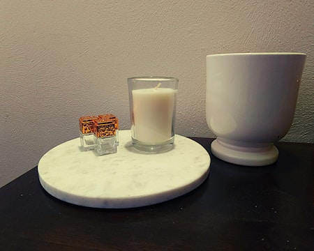 A marble coaster holds a lit white candle, a set of decorative small transparent cubes with orange patterns on top, and is placed next to a large white pot on a dark wooden surface. The background is a plain light-colored wall.