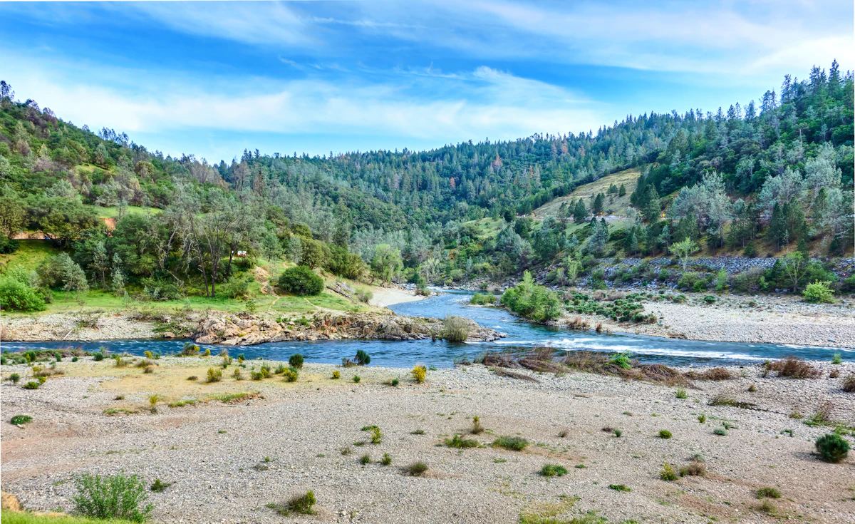 A shallow river and a wide gravel bar under green, wooded hills in the Auburn State Recreation Area