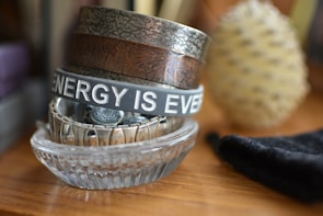 A stack of bracelets and wristbands is neatly arranged in a clear glass dish. The top bracelet is inscribed with the phrase 'ENERGY IS EVERYWHERE'. Various textures and designs are visible on the metal and fabric bands. In the background, a blurred object resembling a pine cone is visible, along with a piece of black cloth.