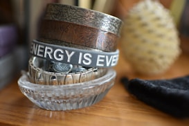 A stack of bracelets and wristbands is neatly arranged in a clear glass dish. The top bracelet is inscribed with the phrase 'ENERGY IS EVERYWHERE'. Various textures and designs are visible on the metal and fabric bands. In the background, a blurred object resembling a pine cone is visible, along with a piece of black cloth.