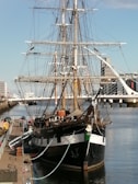 A large, historic sailing ship docked at a harbor, featuring tall masts with rigging and a wooden hull. The ship is surrounded by calm waters, and a modern cityscape with tall buildings and a bridge is visible in the background.
