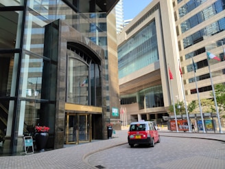 A city scene featuring modern high-rise buildings with large glass windows and a prominent entrance with golden detailing. A red taxi is parked on the curved brick roadway. Several flags on poles are visible, and there are decorative plants in large black pots near the entrance. Pedestrians can be seen walking near the building.
