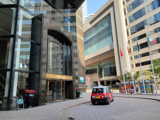 A city scene featuring modern high-rise buildings with large glass windows and a prominent entrance with golden detailing. A red taxi is parked on the curved brick roadway. Several flags on poles are visible, and there are decorative plants in large black pots near the entrance. Pedestrians can be seen walking near the building.