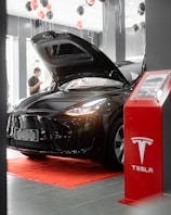 A sleek black car with its hood open is displayed inside a showroom. Red and black balloons float above, and a red platform with a display stand nearby features the Tesla logo. A person stands in the background.