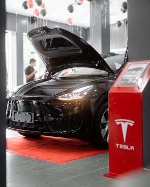 A sleek black car with its hood open is displayed inside a showroom. Red and black balloons float above, and a red platform with a display stand nearby features the Tesla logo. A person stands in the background.