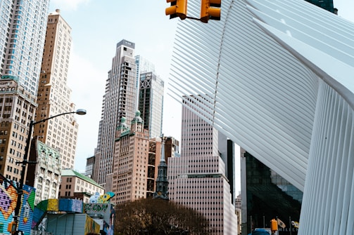Skyscrapers tower over the cityscape, featuring a mix of architectural styles. A modern white structure with vertical slats contrasts with the older, ornate buildings. A traffic light and some colorful street art add urban vibrancy.