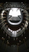 A spiral staircase viewed from below creates a circular frame that draws the eye upward through the center. The architectural design features metallic handrails and dark surfaces, emphasizing the contemporary aesthetic of the space. The lighting is subtle, casting reflections and enhancing the geometric patterns.