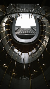 A spiral staircase viewed from below creates a circular frame that draws the eye upward through the center. The architectural design features metallic handrails and dark surfaces, emphasizing the contemporary aesthetic of the space. The lighting is subtle, casting reflections and enhancing the geometric patterns.