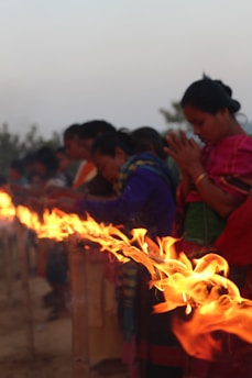 A line of people stand with their heads bowed, engaged in prayer or meditation. In front of them, a long row of flames flickers brightly against a dim backdrop. The individuals are dressed in colorful traditional clothing, suggesting a cultural or religious ceremony.