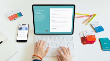 A person is using a tablet with a keyboard on a desk. The tablet displays a screen about search function design and considerations. Nearby, there is a smartphone showing some exploration and creation options. Various colorful cards and pens are scattered on the white surface.