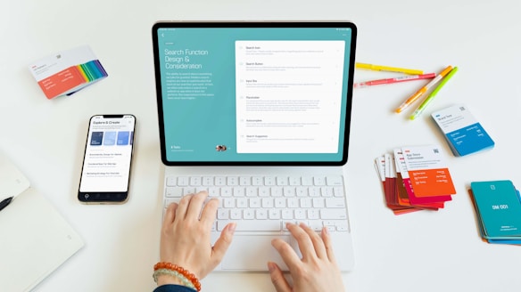 A person is using a tablet with a keyboard on a desk. The tablet displays a screen about search function design and considerations. Nearby, there is a smartphone showing some exploration and creation options. Various colorful cards and pens are scattered on the white surface.
