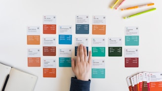 Several colorful cards are laid out on a white surface, arranged in rows. They contain various marketing-related terms and categories. A hand is placed on one of the cards in the center. There are also some pens in different colors placed at the top right corner and an open notebook on the left.