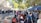A bustling outdoor market scene with numerous stalls and tents shaded by leafy trees. People of various ages, some wearing masks, browse and walk around. Merchandise such as bags and lotions are displayed for sale, and there is a sense of community interaction.