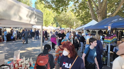 A bustling outdoor market scene with numerous stalls and tents shaded by leafy trees. People of various ages, some wearing masks, browse and walk around. Merchandise such as bags and lotions are displayed for sale, and there is a sense of community interaction.