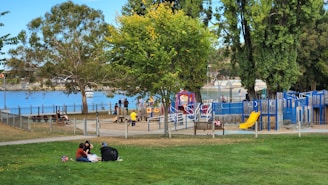 A vibrant park scene by a waterfront with several people enjoying the outdoors. There's a play area with slides and climbing structures, surrounded by trees. A group of three people is having a picnic on the grass in the foreground. Nearby, children are playing and adults are seated on benches. The background features a body of water, with a fence separating the park from the shoreline.