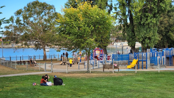 A vibrant park scene by a waterfront with several people enjoying the outdoors. There's a play area with slides and climbing structures, surrounded by trees. A group of three people is having a picnic on the grass in the foreground. Nearby, children are playing and adults are seated on benches. The background features a body of water, with a fence separating the park from the shoreline.