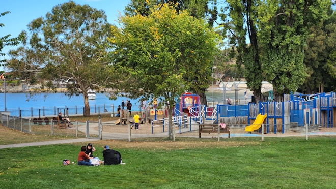 A vibrant park scene by a waterfront with several people enjoying the outdoors. There's a play area with slides and climbing structures, surrounded by trees. A group of three people is having a picnic on the grass in the foreground. Nearby, children are playing and adults are seated on benches. The background features a body of water, with a fence separating the park from the shoreline.