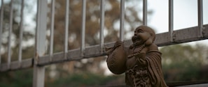 A small, smiling Buddha statue is perched on a wooden shelf in front of a metal railing. The statue holds a sack and displays a joyful expression. The background is a soft, out-of-focus view of trees, suggesting an outdoor setting.