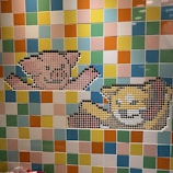 A mosaic wall features two 8-bit style pixel art designs of cartoon animals. The upper design resembles a pink bear with outstretched arms, while the lower depicts a yellow cat with a smiling expression. The background is a colorful arrangement of square tiles in various shades of blue, green, yellow, orange, and pink.