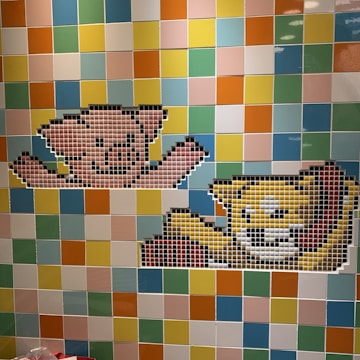 A mosaic wall features two 8-bit style pixel art designs of cartoon animals. The upper design resembles a pink bear with outstretched arms, while the lower depicts a yellow cat with a smiling expression. The background is a colorful arrangement of square tiles in various shades of blue, green, yellow, orange, and pink.