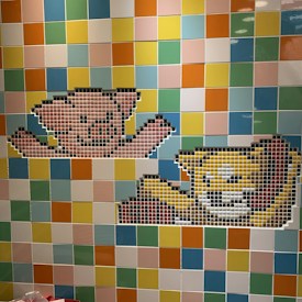 A mosaic wall features two 8-bit style pixel art designs of cartoon animals. The upper design resembles a pink bear with outstretched arms, while the lower depicts a yellow cat with a smiling expression. The background is a colorful arrangement of square tiles in various shades of blue, green, yellow, orange, and pink.