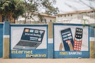 A mural on a concrete wall displays illustrations promoting digital banking services. The left side depicts a laptop with symbols for internet banking, including icons for money, Wi-Fi, and security. The right side shows a hand holding a smartphone with a depiction of mobile banking, showcasing a login screen and a keypad with symbols. The words 'Internet Banking' and 'SMS Banking' are prominently displayed in bold yellow and white letters. The mural is set against a backdrop of trees and buildings.