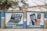 A mural on a concrete wall displays illustrations promoting digital banking services. The left side depicts a laptop with symbols for internet banking, including icons for money, Wi-Fi, and security. The right side shows a hand holding a smartphone with a depiction of mobile banking, showcasing a login screen and a keypad with symbols. The words 'Internet Banking' and 'SMS Banking' are prominently displayed in bold yellow and white letters. The mural is set against a backdrop of trees and buildings.