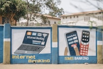 A mural on a concrete wall displays illustrations promoting digital banking services. The left side depicts a laptop with symbols for internet banking, including icons for money, Wi-Fi, and security. The right side shows a hand holding a smartphone with a depiction of mobile banking, showcasing a login screen and a keypad with symbols. The words 'Internet Banking' and 'SMS Banking' are prominently displayed in bold yellow and white letters. The mural is set against a backdrop of trees and buildings.