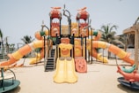 A colorful outdoor playground structure with multiple slides, climbing areas, and a variety of shapes, set on a sandy surface. Surrounding the playground are palm trees and a clear blue sky, creating a tropical setting. The equipment is predominantly orange, yellow, and red, with some green and blue accents.