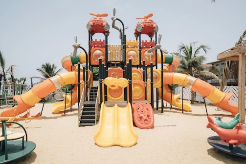 A colorful outdoor playground structure with multiple slides, climbing areas, and a variety of shapes, set on a sandy surface. Surrounding the playground are palm trees and a clear blue sky, creating a tropical setting. The equipment is predominantly orange, yellow, and red, with some green and blue accents.