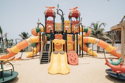 A colorful outdoor playground structure with multiple slides, climbing areas, and a variety of shapes, set on a sandy surface. Surrounding the playground are palm trees and a clear blue sky, creating a tropical setting. The equipment is predominantly orange, yellow, and red, with some green and blue accents.