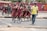 A group of young students in matching maroon school uniforms walk along a roadside accompanied by an adult man in a yellow shirt. They appear to be crossing a street, with some students following behind while others are in the background near a railing and a red mural. The scene seems lively and captures a moment of student life.