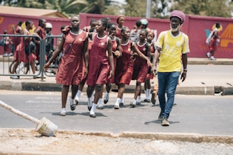 A group of young students in matching maroon school uniforms walk along a roadside accompanied by an adult man in a yellow shirt. They appear to be crossing a street, with some students following behind while others are in the background near a railing and a red mural. The scene seems lively and captures a moment of student life.
