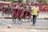 A group of young students in matching maroon school uniforms walk along a roadside accompanied by an adult man in a yellow shirt. They appear to be crossing a street, with some students following behind while others are in the background near a railing and a red mural. The scene seems lively and captures a moment of student life.