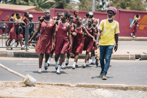 A group of young students in matching maroon school uniforms walk along a roadside accompanied by an adult man in a yellow shirt. They appear to be crossing a street, with some students following behind while others are in the background near a railing and a red mural. The scene seems lively and captures a moment of student life.