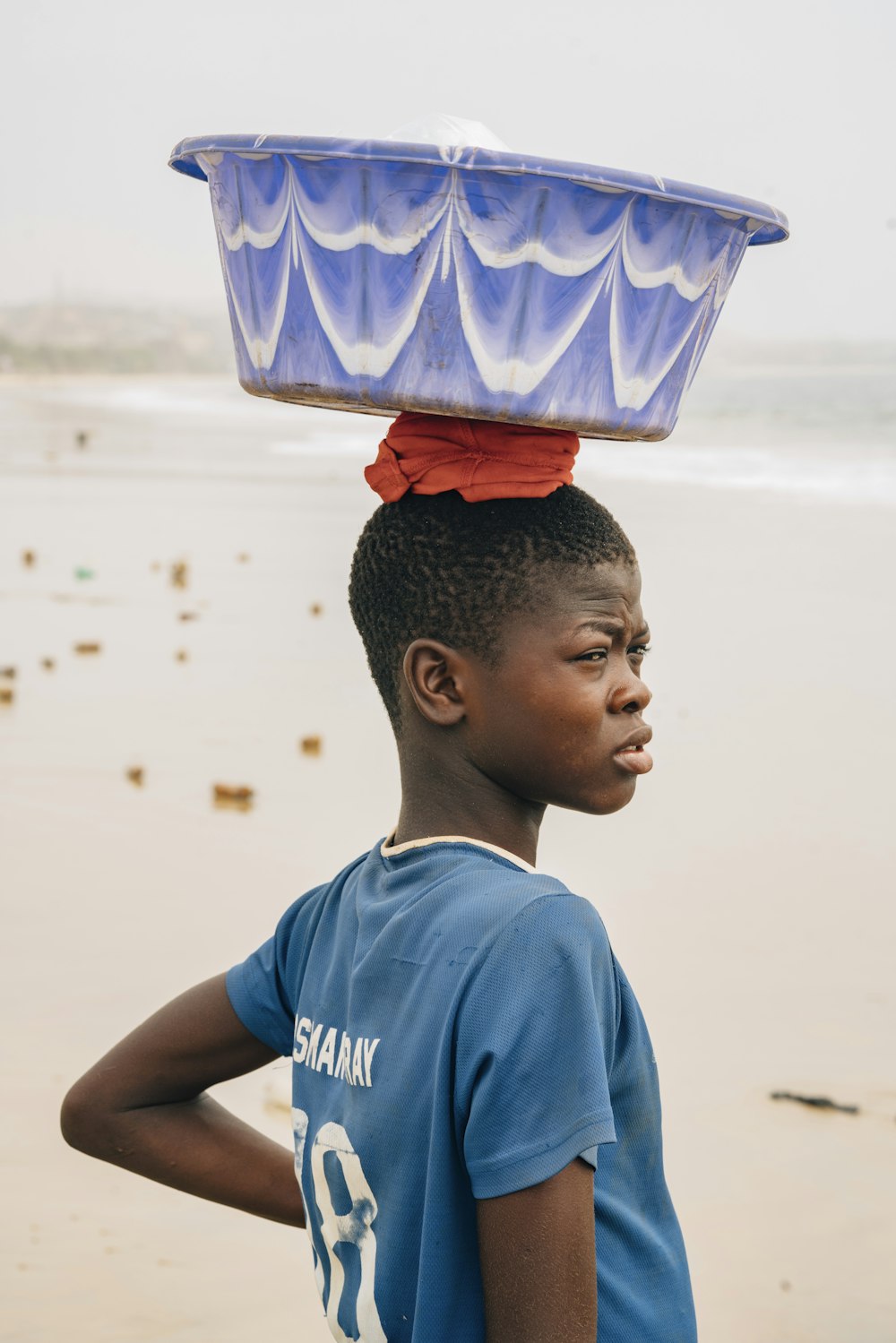 um menino de pé na praia com um balde na cabeça