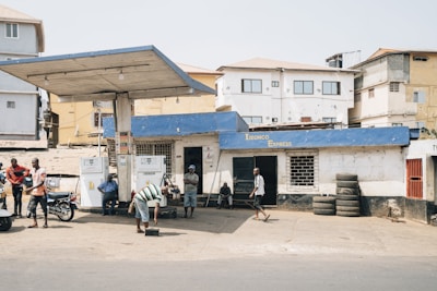A small, local gas station with a blue roof and white walls, including two fuel pumps at the front. People are casually standing or sitting around, with a few motorcycles parked nearby. Several used tires are stacked against the wall, and the backdrop features residential buildings.