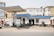 A small, local gas station with a blue roof and white walls, including two fuel pumps at the front. People are casually standing or sitting around, with a few motorcycles parked nearby. Several used tires are stacked against the wall, and the backdrop features residential buildings.