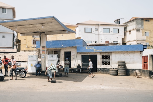 A small, local gas station with a blue roof and white walls, including two fuel pumps at the front. People are casually standing or sitting around, with a few motorcycles parked nearby. Several used tires are stacked against the wall, and the backdrop features residential buildings.