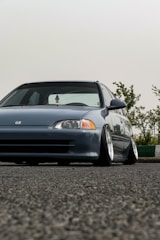 A low-angle view of a modified gray car with lowered suspension, positioned on an asphalt surface. The car has a sleek design with prominent wheel rims and a clear front view. In the background, there are green shrubs against a pale sky.