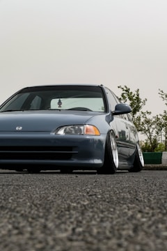 A low-angle view of a modified gray car with lowered suspension, positioned on an asphalt surface. The car has a sleek design with prominent wheel rims and a clear front view. In the background, there are green shrubs against a pale sky.