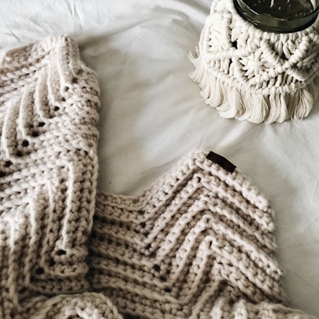 A cozy, knitted blanket with a zigzag pattern is laid out alongside a macrame plant hanger, both positioned on a soft, white fabric surface. The textures are rich and tactile, highlighting intricate craftsmanship.