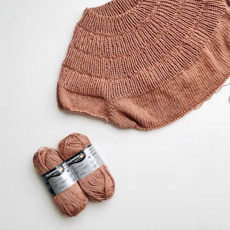 A knitted piece of fabric in a warm, earthy brown hue is laid out on a white surface. Next to it, two skeins of yarn wrapped with labels indicating 'Fashion Soft Linen Mix' are positioned.