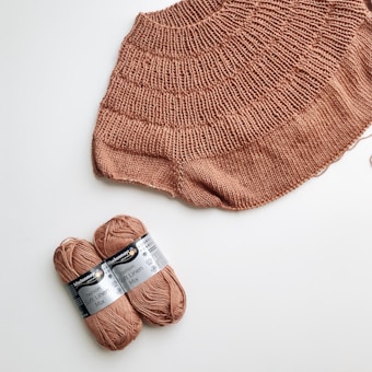 A knitted piece of fabric in a warm, earthy brown hue is laid out on a white surface. Next to it, two skeins of yarn wrapped with labels indicating 'Fashion Soft Linen Mix' are positioned.