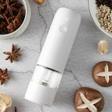 A sleek, modern white grinder is placed on a textured gray surface surrounded by an assortment of spices and garlic. Star anise and peppercorns are scattered around, alongside whole garlic bulbs and mushrooms with cross marks.