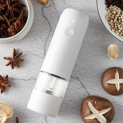 A sleek, modern white grinder is placed on a textured gray surface surrounded by an assortment of spices and garlic. Star anise and peppercorns are scattered around, alongside whole garlic bulbs and mushrooms with cross marks.