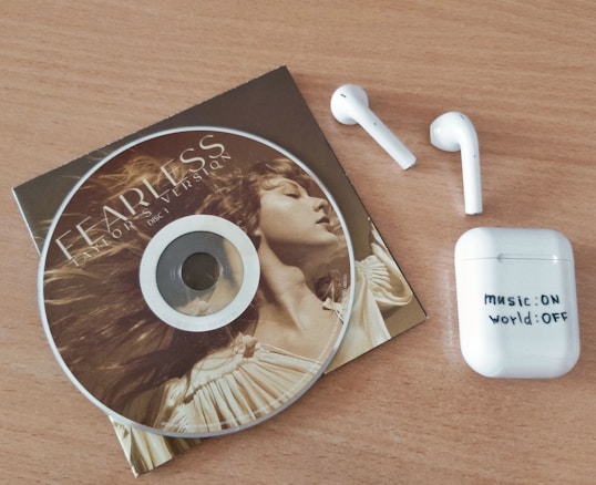 A CD with art featuring a person and text on it is placed on a wooden surface. Next to it are a pair of wireless earbuds and a charging case labeled with 'music: ON world: OFF'.