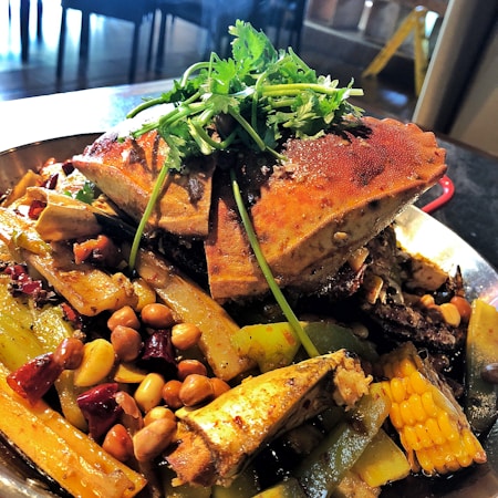 A large, well-prepared seafood dish featuring a grilled crab as the main component, accompanied by a mix of vegetables including corn, green peppers, and whole peanuts. The dish is garnished with fresh cilantro and drizzled with a rich, savory sauce. Vibrant colors and textures are highlighted by the lighting, creating an appetizing presentation.