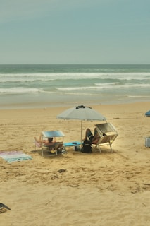 A sandy beach with two people lounging on deck chairs under umbrellas. The ocean waves gently roll in the background, creating a serene and relaxing atmosphere. Various beach towels are spread out nearby.